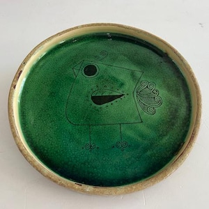 May include: A round ceramic plate with a green center and a beige rim. The plate features a whimsical black line drawing of a bird. The bird has a square body, a round eye, and a detailed wing.