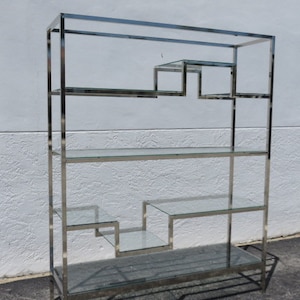 May include: A tall, chrome-finished shelving unit with multiple glass shelves. The design includes a series of stepped shelves, adding visual interest. The unit is free-standing and suitable for displaying items.
