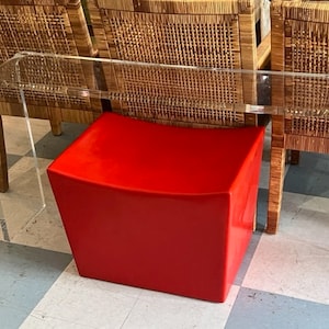Könnte beinhalten: Ein klarer Konsolentisch aus Acryl mit geschwungener Oberseite und Beinen. Ein leuchtend roter, würfelförmiger Hocker befindet sich unter dem Tisch. Korbstühle sind im Hintergrund.