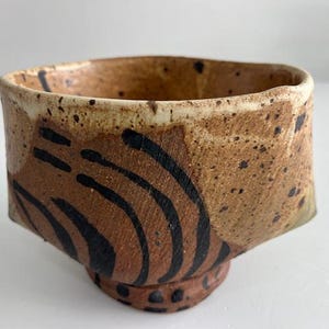 Puede incluir: Un cuenco de cerámica hecho a mano con un diseño único. El cuenco presenta una forma cuadrada con esquinas redondeadas, un exterior marrón moteado y líneas negras curvas. La base es un pedestal más pequeño y redondeado. El borde es de color más claro.