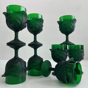 May include: A collection of green-tinted glass goblets. The goblets feature a leaf-like design around the bowl and stem. Some are upright, while others are inverted or lying on their sides, creating a visually interesting arrangement.