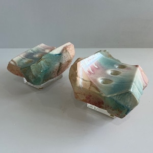 Peut inclure: Deux porte-savons en céramique avec un extérieur rugueux rappelant la roche et un intérieur lisse émaillé. L'émail présente un mélange de couleurs bleu sarcelle, rose et crème. Le porte-savon de droite a trois trous. Chaque pièce repose sur un socle.