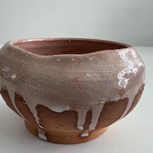 Puede incluir: Un cuenco de cerámica hecho a mano, de forma redondeada y con una amplia abertura. El cuenco es de color terracota cálido, con un esmalte texturizado blanco-rosado claro que gotea por los lados. Se apoya sobre una pequeña base circular.