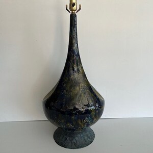 May include: A tall, teardrop-shaped ceramic table lamp base with a glossy, mottled glaze in shades of deep navy blue, forest green, and metallic gold. The lamp features a narrow neck, a wide rounded body, and a circular base with a brass socket.
