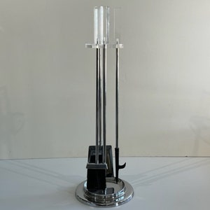 Puede incluir: Un juego de herramientas para chimenea cromado con un soporte de acrílico transparente. El juego incluye un atizador, una pala y un cepillo. La base tiene un diseño circular con un acabado pulido. Las herramientas están dispuestas verticalmente.