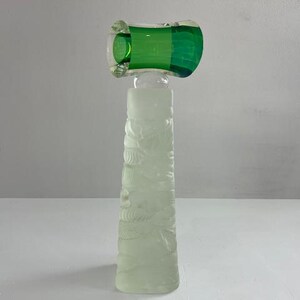 Könnte beinhalten: Eine hohe, dekorative Glasflasche mit einem mattierten, geschnitzten Körper und einem leuchtend grünen Verschluss. Der Verschluss hat eine rechteckige Form mit abgerundeten Kanten. Der Flaschenkörper weist ein geschnitztes Blumenmuster auf.