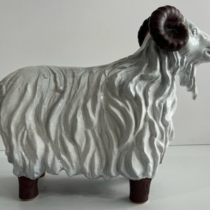 Op de afbeelding: Een keramieken ram-beeldje met een glanzende witte afwerking en diepe, vloeiende gebeeldhouwde texturen die dikke wol voorstellen. De ram heeft donkerbruine, gekrulde hoorns en bijpassende donkerbruine, cilindervormige poten in zijaanzicht.