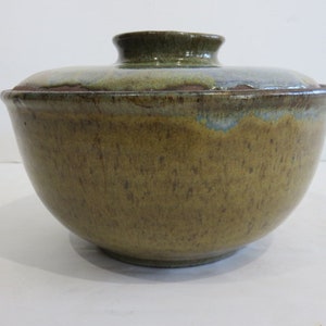 Może przedstawiać: Ceramiczna miska z dopasowaną pokrywką, pokryta nakrapianą oliwkowozieloną i brązową glazurą. Zewnętrzna powierzchnia ma cętkowaną fakturę, podczas gdy pokrywka wykazuje gładkie, błyszczące wykończenie z subtelnymi niebieskimi i zielonymi odcieniami.