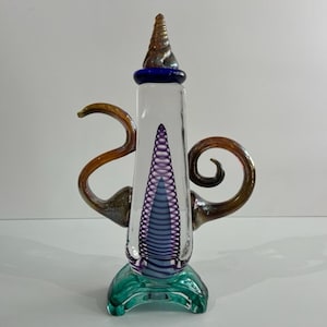 Puede incluir: Una tetera decorativa de cristal transparente con un diseño en espiral en el interior. La tetera tiene asas marrones, un borde azul y una parte superior marrón en espiral. Se apoya sobre una base verde.