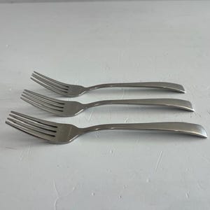May include: A set of three stainless steel forks arranged in a parallel stack. Each fork features a smooth, polished finish with four tines and a slightly curved handle that tapers toward the end, showcasing a clean and simple design.