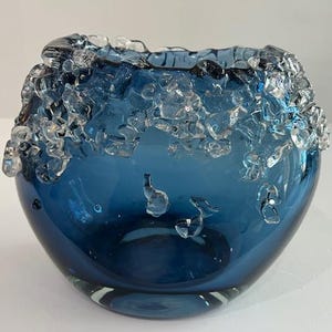 Könnte beinhalten: Eine runde, blaue Glasschale mit einem strukturierten, klaren Glasrand. Die Schale hat eine glatte, glänzende Oberfläche und weist dekorative, tropfenförmige Glaselemente im Inneren auf. Ein einzigartiges Dekorationsstück.