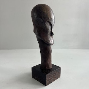Könnte beinhalten: Eine dunkelbraune, glatte, abstrakte Skulptur in Form eines Kopfes und Halses, die auf einem quadratischen, dunkel gemaserten Holzsockel montiert ist. Die Skulptur hat eine abgerundete Oberseite, einen schmalen Hals und ein subtil vertieftes Gesichtsprofil.