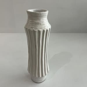 May include: A tall, off-white ceramic vase featuring a vertical ribbed texture along its body. The vase has a flared neck and a slightly tapered base, with subtle greyish-brown horizontal brushstroke details visible across the surface.