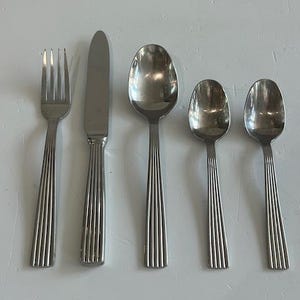 May include: A five-piece stainless steel flatware set consisting of a fork, a dinner knife, a large spoon, and two smaller spoons. Each piece features a polished finish and a handle with vertical ribbed detailing running along the length.