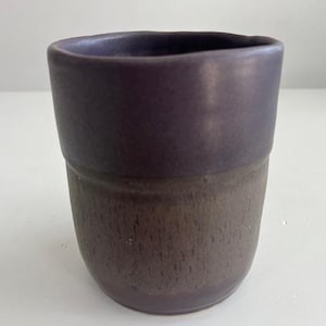 Könnte beinhalten: Handgefertigte Keramiktasse mit zweifarbigem Design. Der obere Teil ist ein tiefes Lila, während der untere Teil eine strukturierte, erdartige braune Optik hat. Die Tasse hat einen abgerundeten Boden und einen glatten, leicht ausgestellten Rand.