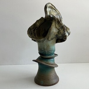 Puede incluir: Una escultura de cerámica abstracta con una forma orgánica texturizada. La escultura presenta una base marrón oscuro que transiciona a tonos turquesa y marrón claro. La parte superior tiene una forma grande y abierta en forma de concha.