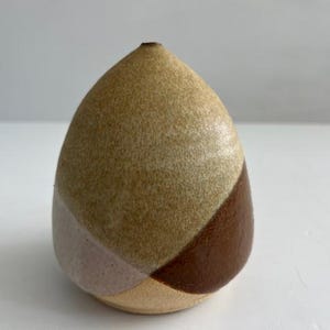 Puede incluir: Un jarrón de cerámica hecho a mano con forma de lágrima. El jarrón presenta un cuerpo moteado de color marrón claro con secciones geométricas de color rosa claro, marrón claro y marrón oscuro. Tiene una pequeña abertura en la parte superior y es adecuado para flores secas o como pieza decorativa.