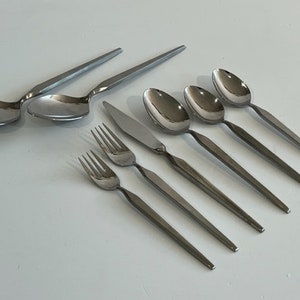 May include: A set of stainless steel flatware, including spoons, forks, and knives. The silverware has a sleek, modern design with long, slender handles and a polished finish. The spoons have a rounded bowl shape.