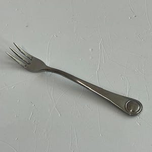 May include: A stainless steel fork with three tines and a long, slender handle. The handle has a rounded end with a circular design. The fork is set against a white textured background.