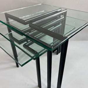 May include: A set of three nesting tables featuring clear rectangular glass tops and black metal frames. Each table has four straight cylindrical legs and a tiered design, allowing the smaller tables to slide underneath the largest one.