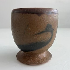 Puede incluir: Una pequeña taza de cerámica hecha a mano con cuerpo redondeado y base con pie. La taza es de color marrón moteado con un borde marrón oscuro y un diseño abstracto negro. La taza mide aproximadamente 7.6 cm de alto.