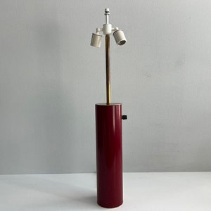 Peut inclure: Une base de lampe de table composée d'un corps cylindrique haut d'un rouge profond avec une finition lisse. Une fine tige métallique couleur laiton s'élève du centre, surmontée d'une douille double blanche et d'un petit interrupteur rotatif noir.