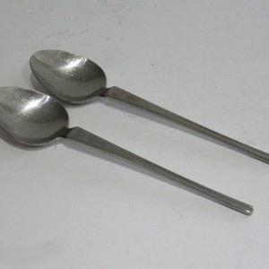 May include: Two stainless steel spoons with oval bowls and flat, rectangular handles. The spoons are positioned diagonally on a white surface, with one slightly overlapping the other. The spoons have a shiny, reflective surface.