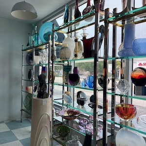 May include: A display of glass art pieces on chrome shelves. The collection includes vases, bowls, and decorative objects in various colors, shapes, and sizes. The shelves are arranged in front of a window.