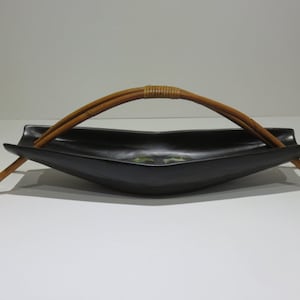 Könnte beinhalten: Eine mattschwarze, längliche, bootsförmige Keramikschale mit einem zentralen, grün und braun bemalten Design. Die Schale ist mit einem geschwungenen, hellbraunen Bambusgriff versehen, der zu beiden Seiten herausragt und mit Rattan umwickelt ist.