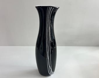 Puede incluir: Un jarrón de cristal alto y esbelto con un acabado negro brillante. El jarrón presenta un borde acampanado y una serie de finas líneas blancas verticales que recorren el lateral, creando un marcado contraste con la superficie oscura y reflectante.