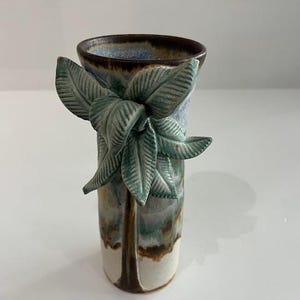 Puede incluir: Un jarrón de cerámica de forma cilíndrica, con un grupo de hojas verdes en el lateral. El jarrón tiene un degradado de color, que va del marrón al blanco, con toques de azul y verde. El borde es marrón oscuro.