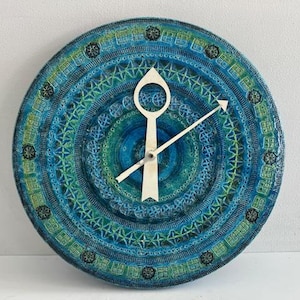 May include: A round wall clock featuring a textured, multi-layered surface in shades of blue and green. The clock face displays intricate, repeating geometric patterns and small dark circular accents. Two light-colored, pointed hands are centered.