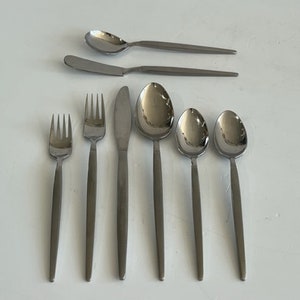 May include: A set of eight stainless steel flatware pieces arranged in a row and two stacked horizontally. The collection includes two forks, two knives, and four spoons, all featuring a polished metallic finish with slender, tapered handles.
