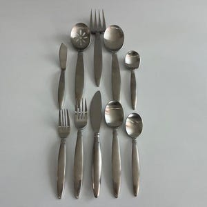 May include: A collection of ten stainless steel flatware pieces arranged in two columns, including forks, spoons, a slotted spoon, and knives. The set features a brushed metal finish with smooth, tapered handles and rounded edges throughout.