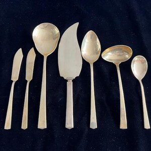 May include: A collection of silver-colored serving utensils, including butter knives, spoons, a cake server, and a ladle, arranged on a dark blue surface. The utensils have long, slender handles and are designed for serving food.