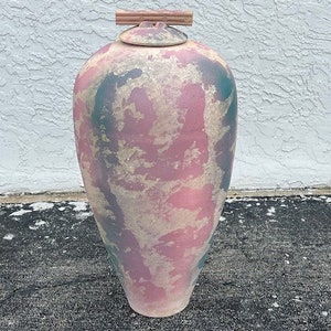 May include: A large, decorative ceramic urn with a lid. The urn is painted with a mottled pattern of pink, teal, and beige. The lid has a small handle made of wood. The urn is tall and has a rounded shape.