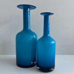 May include: Two blue glass vases of different sizes. The vases have a bottle-like shape with a wide base, a long, slender neck, and a flared top. The color is a vibrant, translucent blue, with a darker shade at the base. The vases are likely decorative items.