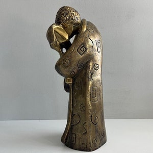 May include: A bronze sculpture of two figures in an embrace. The figures are closely intertwined, with detailed patterns on their robes. The sculpture has a warm, metallic finish and is placed on a white surface. The theme is love and connection.
