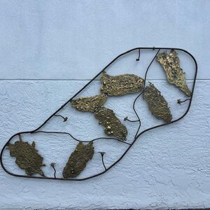 May include: A wall-mounted metal sculpture featuring a dark, thin, irregular wire frame that encloses several textured, gold-toned brass plates with organic, pitted surfaces and small, delicate wire stems with tiny buds attached to the frame.