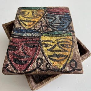 Puede incluir: Una baldosa rectangular de cerámica marrón texturizada con cuatro caras estilizadas y coloridas. Cada cara está delineada en negro y presenta una combinación de colores diferente: amarillo, rojo, azul y verde. La baldosa descansa sobre una bandeja marrón a juego.