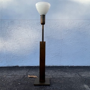 May include: A floor lamp featuring a tall, rectangular bronze-toned metal base and a slender central pole. The lamp is topped with a white, bowl-shaped glass shade that has subtle horizontal ridges along its exterior surface.