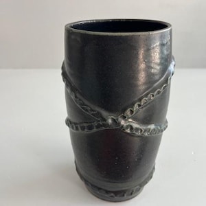 Pode incluir: Um vaso cilíndrico de cerâmica com acabamento em preto carvão escuro. A superfície exterior apresenta um padrão em relevo com textura de corda que envolve o corpo num design entrecruzado, criando uma aparência tátil e dimensional.