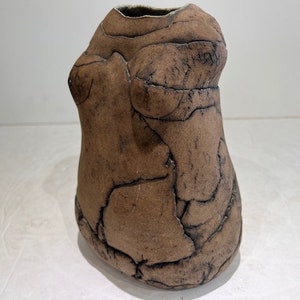 Puede incluir: Un jarrón de cerámica marrón con forma de torso, con una superficie rugosa y texturizada. El jarrón tiene un aspecto agrietado con líneas oscuras y un interior liso y oscuro. El diseño recuerda a la cerámica antigua.
