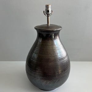 May include: A dark brown ceramic table lamp with a textured surface. The lamp has a bulb socket and a metal stem. The lamp's body is a rounded shape, and the lamp is sitting on a white surface.