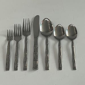 May include: A stainless steel flatware set with a bamboo-style handle design. The set includes forks, knives, and spoons. The utensils are arranged on a white surface, showcasing their polished, reflective surfaces.