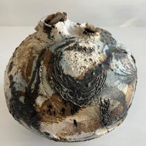 Puede incluir: Un jarrón de cerámica redondo con una superficie texturizada e irregular que presenta una paleta de colores moteados en crema, marrón oscuro y azul apagado. El recipiente tiene una pequeña abertura irregular en la parte superior.