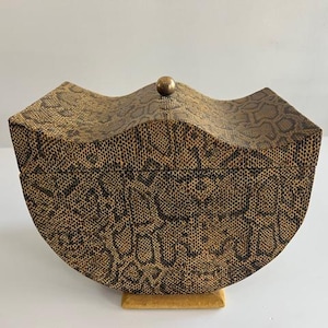 Puede incluir: Una caja semicircular con un patrón texturizado de piel de serpiente en marrón y negro. La tapa tiene un borde superior curvo en forma de onda y está rematada con un pequeño pomo esférico de color latón centrado en la parte superior.