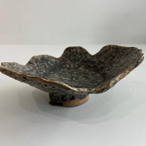 Puede incluir: Un cuenco de cerámica hecho a mano, moteado, con una forma orgánica. El cuenco presenta una superficie oscura y moteada y una base elevada. El borde tiene un borde texturizado e irregular. Adecuado para servir o exhibir. Diámetro aproximado de 15 cm.