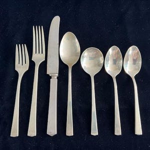 May include: A set of silver-colored flatware, including forks, a knife, and spoons, arranged on a dark blue surface. The utensils have a simple, elegant design, suitable for formal dining. The set includes two forks, a knife, and four spoons.
