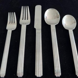 May include: A set of five silver-colored flatware pieces, including two forks, a knife, and two spoons. Each piece has a ribbed design on the handle. The flatware is arranged on a black surface.